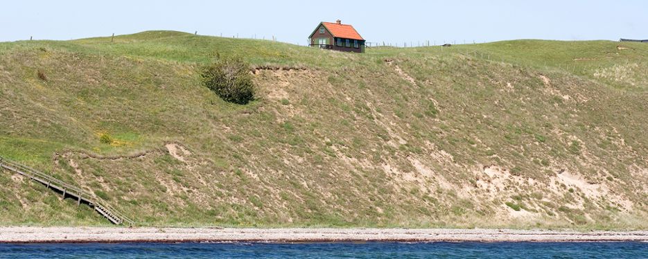 Litet hus högst upp på en kulle med en hög sandsluttning ner mot havet.
