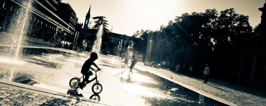 Litet barn cyklar i en fontän. Motljus.