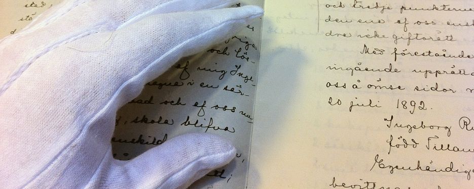 En handskbeklädd hand bläddrar försiktigt i en gammal bok med handskriven text.